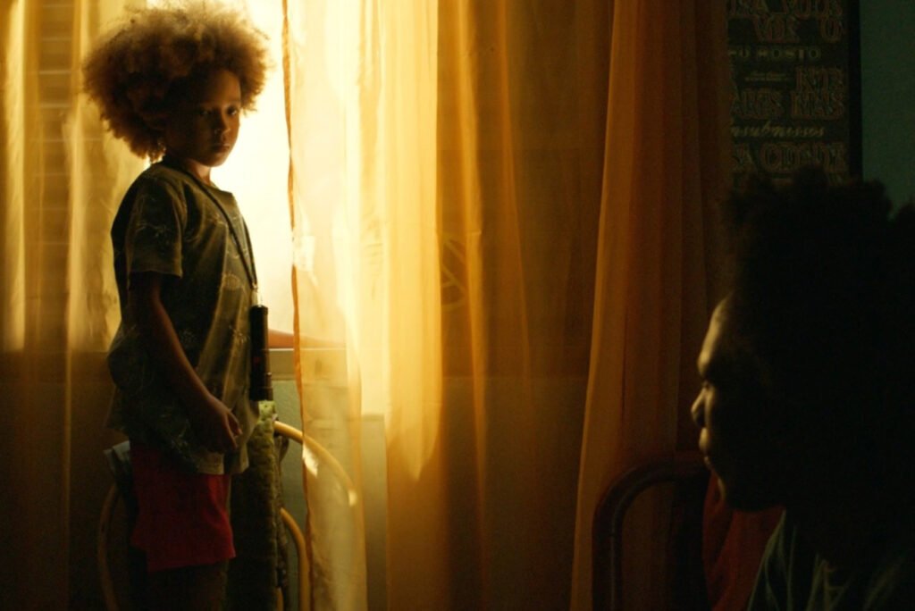 Efraim Santos como Tutu aparece em pé diante de uma janela iluminada por luz dourada, com expressão séria, enquanto outra pessoa surge em silhueta no canto direito, em cena do filme ‘Nosso Segredo’.
