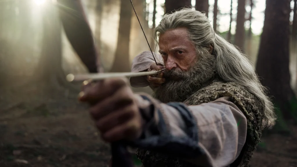 Hugh Jackman caracterizado como Robin Hood, com cabelos longos e barba grisalha, aponta um arco e flecha em meio a uma floresta iluminada pela luz do sol em cena de ‘A Morte de Robin Hood’.