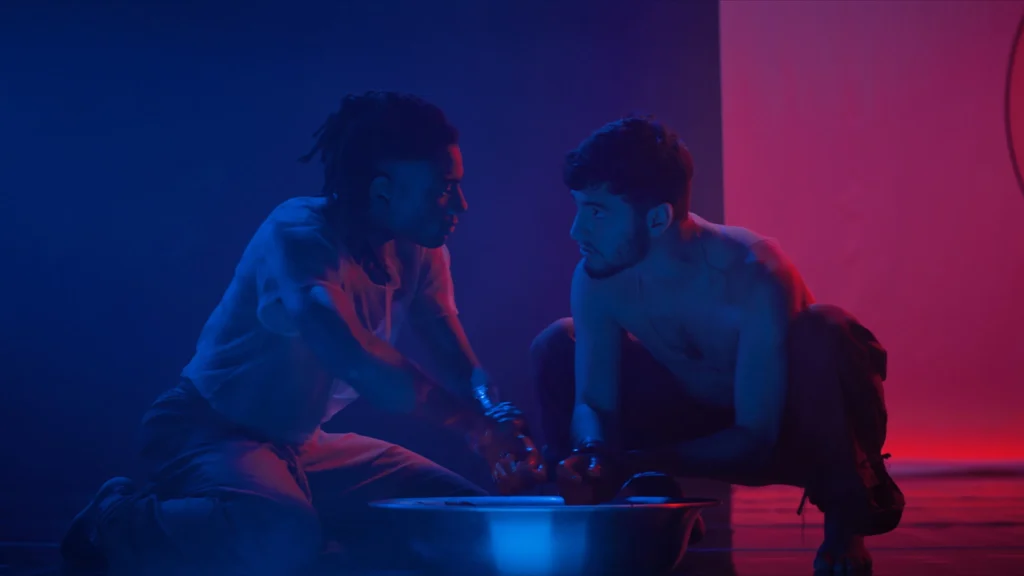 Gabriel Faryas como Matias e Cirillo Luna como Rafael aparecem frente a frente em um ambiente iluminado por luzes azul e vermelha, em uma cena de tensão e proximidade do filme ‘Ato Noturno’.