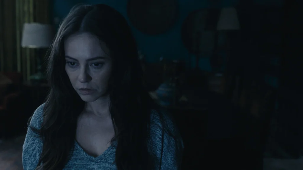 Paolla Oliveira como Ana aparece em primeiro plano com expressão tensa e olhar fixo, em um ambiente escuro iluminado por tons azulados, em cena do filme ‘Herança de Narcisa’.