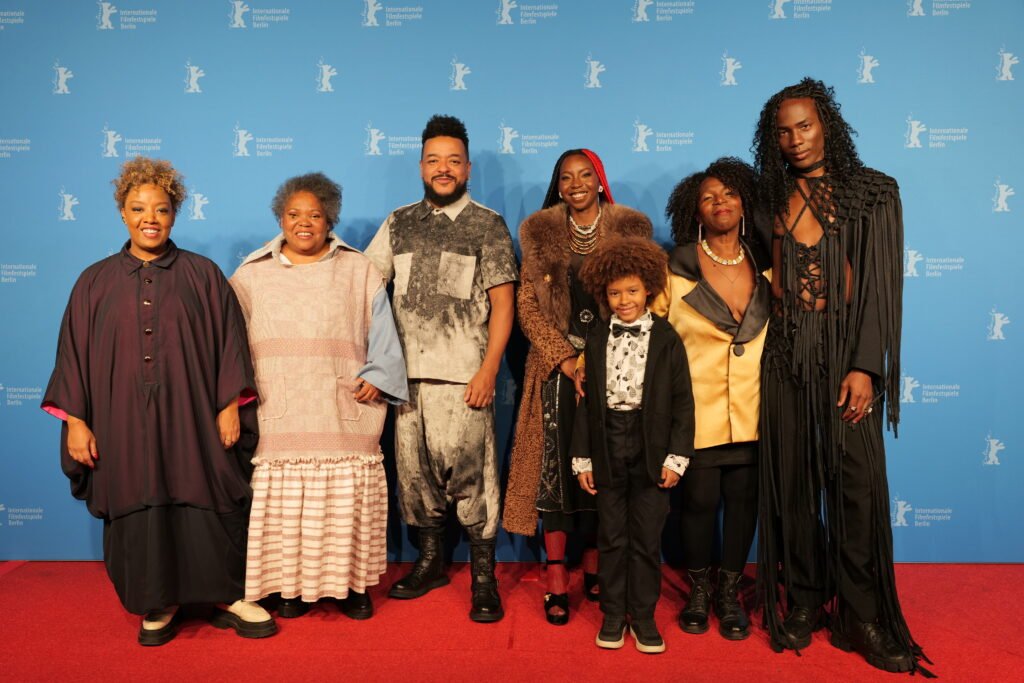 Grace Passô posa ao lado de Robert Frank, Efraim Santos, Jéssica Gaspar, Flip, Ju Colombo e Marisa Revert, integrantes do elenco de ‘Nosso Segredo’, no tapete vermelho do Festival Internacional de Cinema de Berlim. O grupo aparece diante do painel azul da Berlinale durante a apresentação do filme no festival.