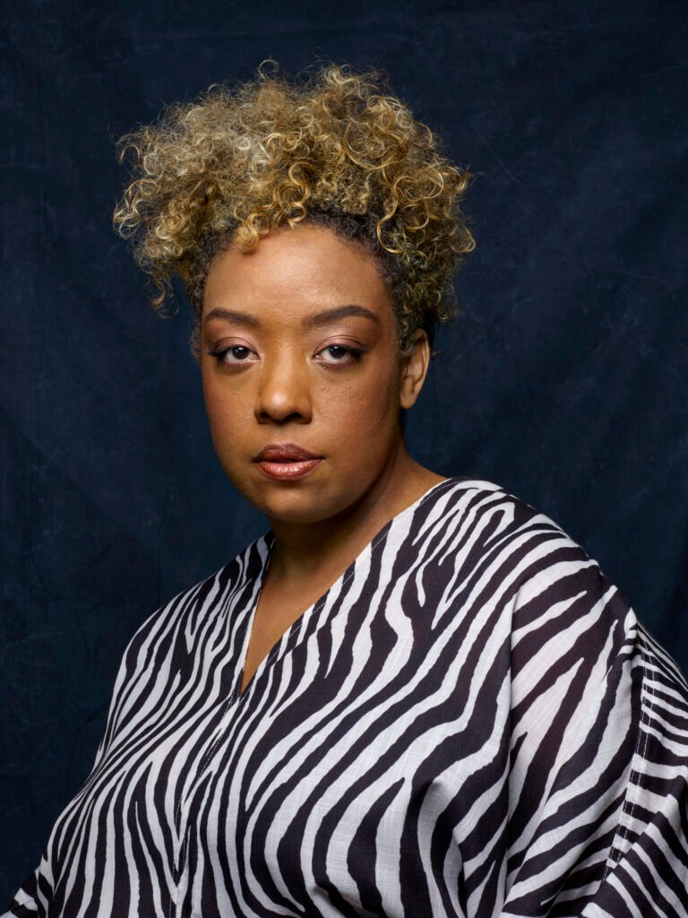 Retrato da diretora e roteirista Grace Passô, responsável por ‘Nosso Segredo’, posando diante de um fundo azul-escuro. Ela aparece com expressão séria, cabelos cacheados presos no alto e veste uma blusa preta e branca com estampa de zebra.