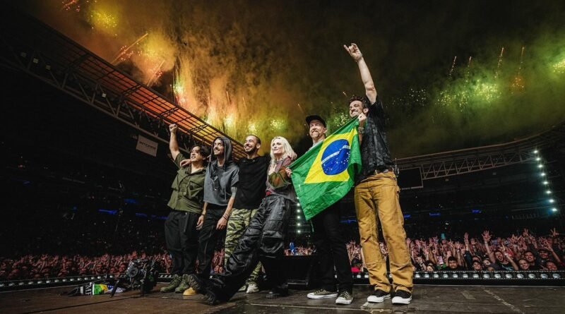 A banda Linkin Park aparece reunida no palco do Allianz Parque, em São Paulo, após o show de 15 de novembro de 2024. Da esquerda para a direita estão Joe Hahn, Colin Brittain, Mike Shinoda, Emily Armstrong, Dave “Phoenix” Farrell e o guitarrista de turnê Alex Feder. O grupo posa diante da multidão, segurando uma bandeira do Brasil, enquanto fogos de artifício verdes e amarelos iluminam o estádio ao fundo. A imagem é usada como capa da crítica do documentário ‘LINKIN PARK - UNSHATTER’ no blog Multi Nerdz.