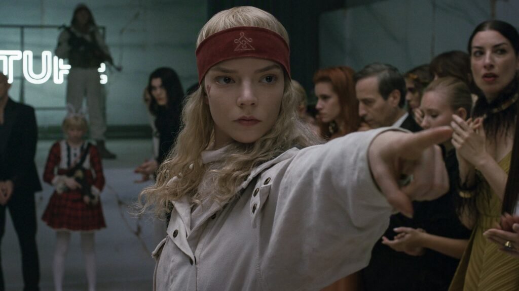 Anya Taylor-Joy em cena do filme Sacrifício, usando faixa vermelha na cabeça e roupa clara, com o braço estendido para a frente em um ambiente interno cercado por outras pessoas ao fundo. A atriz aparece em destaque no centro da imagem, utilizada como ilustração no blog Multi Nerdz.