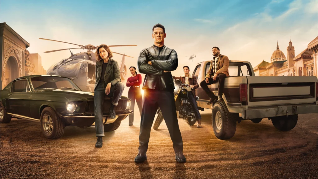 Imagem promocional de ‘Matchbox – O Filme’, da Apple TV, usada no blog Multi Nerdz. A arte reúne Jessica Biel como Aimee, Teyonah Parris como Charmaine, John Cena como Sean, Arturo Castro como Ted e Sam Richardson como Crosby em meio a carros, motocicleta e helicópteros. John Cena aparece em destaque no centro, de braços cruzados, enquanto o grupo posa diante de uma paisagem ensolarada com arquitetura monumental ao fundo, reforçando o clima de ação e aventura do filme.