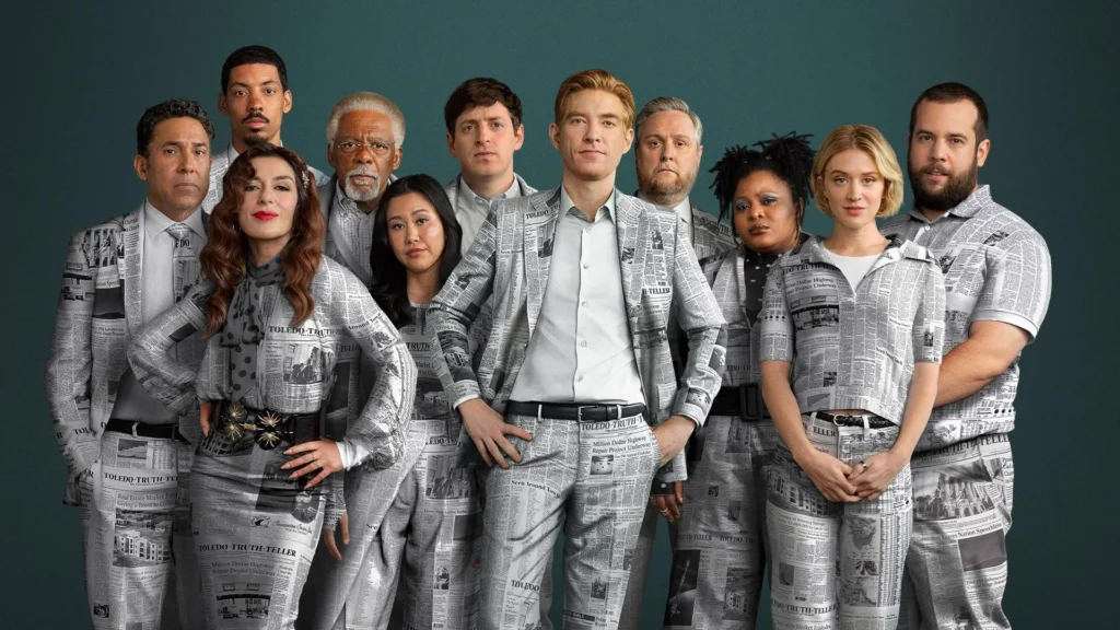 Imagem promocional da 2ª temporada de ‘The Paper’ usada no blog Multi Nerdz, reunindo o elenco principal diante de um fundo verde-azulado. Domhnall Gleeson, Sabrina Impacciatore, Chelsea Frei, Melvin Gregg, Gbemisola Ikumelo, Tim Key, Ramona Young, Eric Rahill, Duane Shepard Sr., Alex Edelman e Oscar Nuñez aparecem juntos usando figurinos estampados com páginas do jornal fictício Toledo Truth Teller.